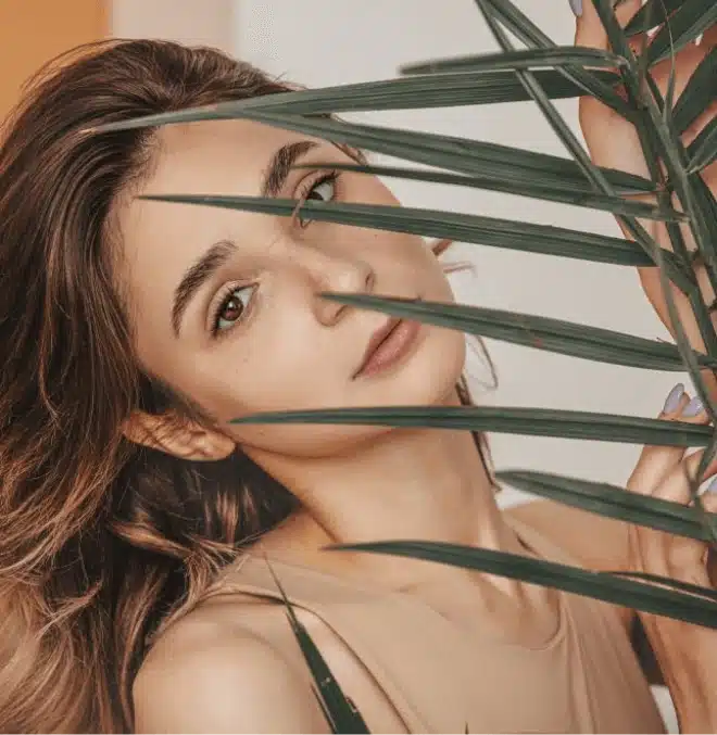 A woman with long, wavy hair peers through green palm leaves, creating a serene and natural vibe. She wears a neutral top and has a calm expression.